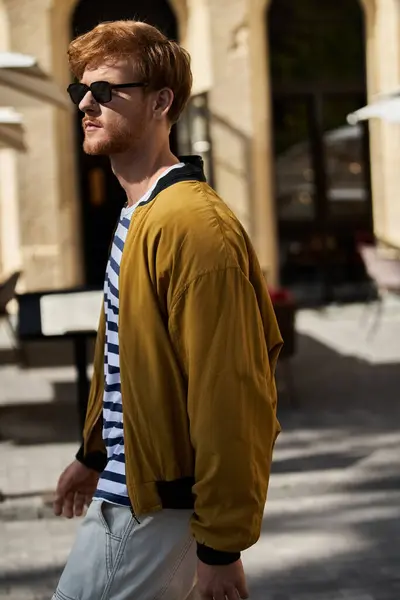 Модный молодой человек с рыжими волосами и модными солнцезащитными очками уверенно прогуливается по городской улице. — стоковое фото
