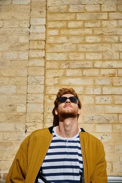 Joven pelirrojo en traje de debonair se para con confianza frente a una pared de ladrillo con gafas de sol. - foto de stock