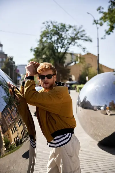 A debonair young man with red hair stands next to a shiny metal ball in the city. — Stock Photo