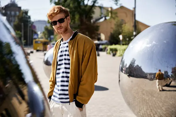 A stylish red-haired man stands confidently in front of a massive metallic sphere. — Stock Photo