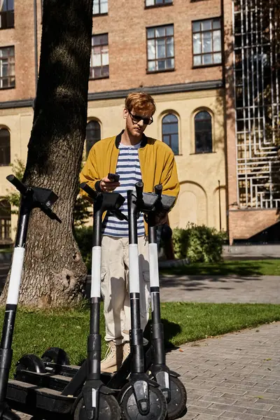Young red-haired man in debonair attire standing by scooters on a city sidewalk. — Stock Photo