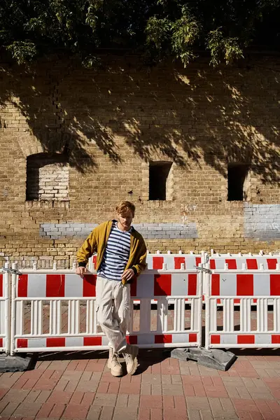 A stylish young man with red hair standing elegantly next to a white and red fence in the city. — Stock Photo