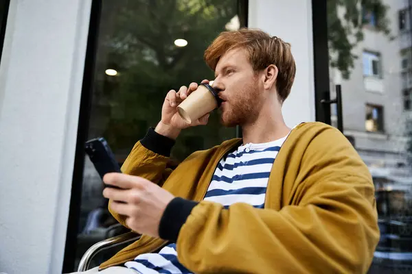 Junger rothaariger Mann in Debonair-Kleidung sitzt da, schreibt SMS aufs Handy und trinkt Kaffee. — Stockfoto