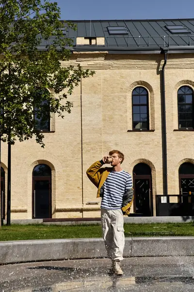 Young man with red hair in stylish attire drinking coffee in front of urban building. — Stock Photo