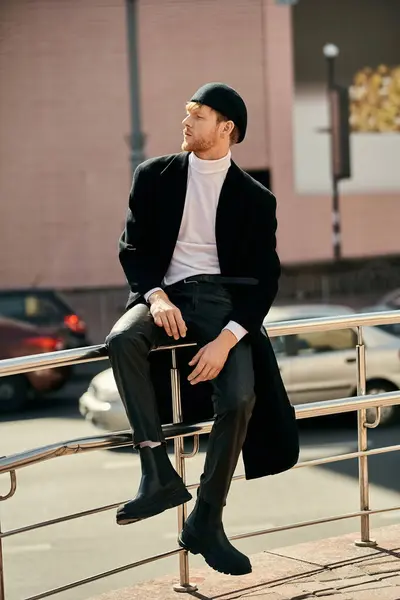 A stylish young man with red hair, sporting a black coat and hat, is elegantly seated on a rail in the city. — Stock Photo