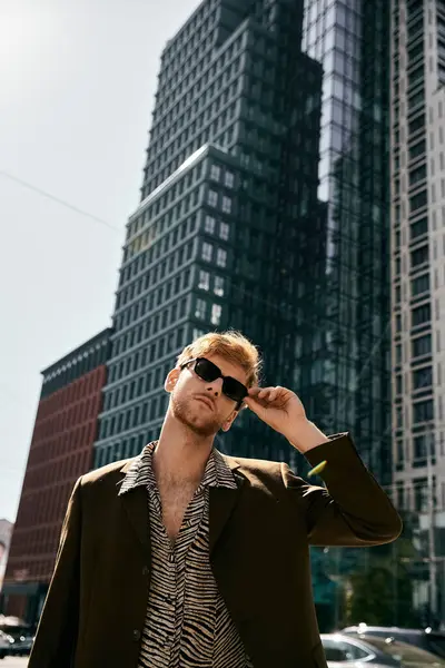 Un hombre pelirrojo con traje y gafas de sol se para con confianza frente a edificios altos. - foto de stock