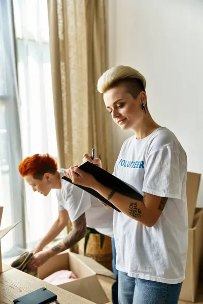 A young lesbian couple in matching volunteer t-shirts organizing boxes in a room for charity work. — Stock Photo