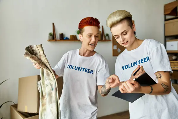 Pareja joven lesbiana en camisetas voluntarias, de pie al lado del otro, comprometida en obras de caridad. - foto de stock