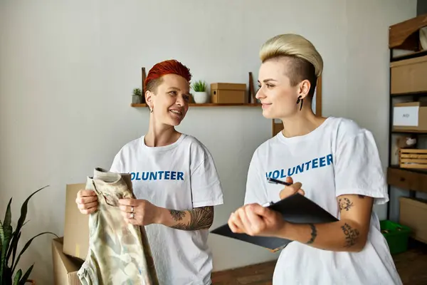 Young lesbian couple in volunteer t-shirts stand together in a room, engaged in charity work. — Stock Photo