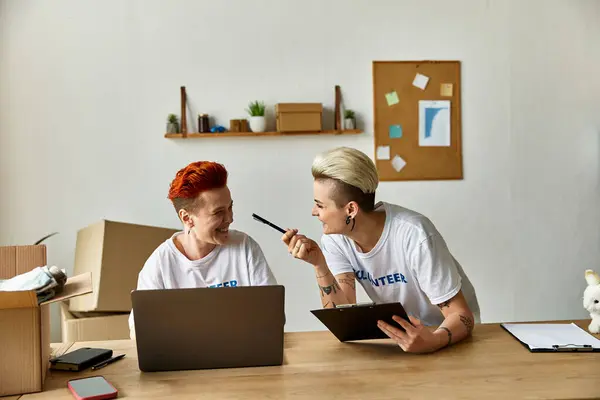 Dos jóvenes mujeres LGBTQ con camisetas voluntarias trabajan juntas en computadoras portátiles en una mesa. - foto de stock