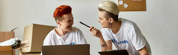 A young lesbian couple, clad in volunteer t-shirts, are deeply focused as they work on their laptop together. — Stock Photo