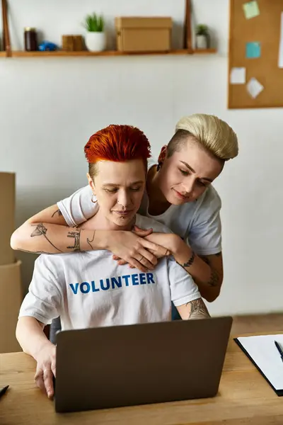 Jeune couple de lesbiennes en t-shirts bénévoles travaillant ensemble sur un ordinateur portable pour un projet de charité. — Photo de stock