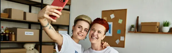 Una mujer entusiasta felizmente toma una selfie con su amiga, ambas usando camisetas voluntarias. - foto de stock
