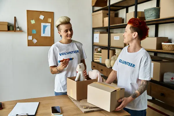 Jeune couple de lesbiennes en t-shirts bénévoles debout dans la chambre avec des boîtes, incarnant la générosité et la compagnie. — Photo de stock
