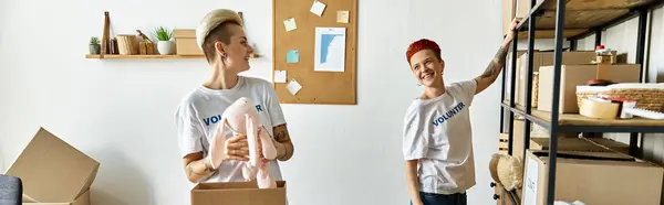 A young lesbian couple in volunteer shirts, standing in a room filled with boxes, working together for charity. — Stock Photo