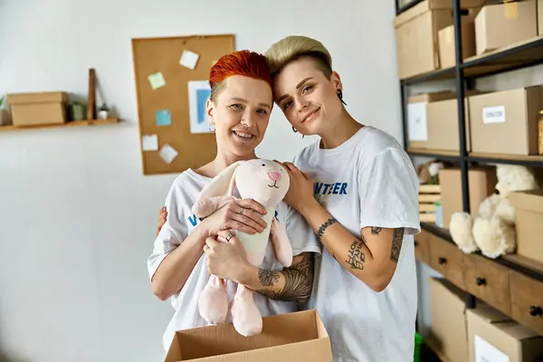 Young lesbian couple in volunteer t-shirts holding a stuffed animal, spreading joy during charity work. — Stock Photo