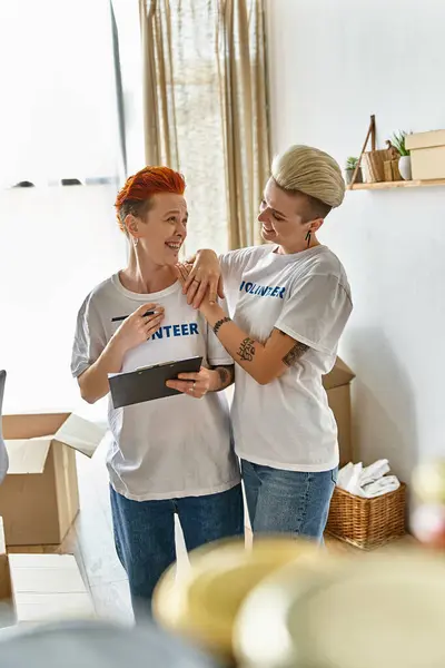 A lesbian couple in volunteer t-shirts, standing side by side, actively engaging in charity work together. — Stock Photo
