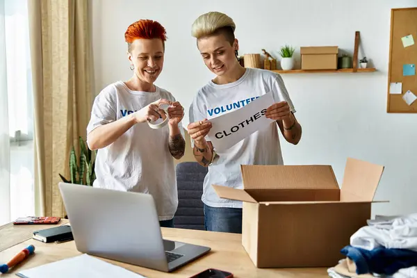 Young lesbian couple in volunteer t-shirts standing in front of a laptop, working together for a charity cause. — Stock Photo