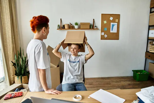 A woman, part of a young lesbian couple, sits on a desk with a box on her head while volunteering in charity work. — Stock Photo