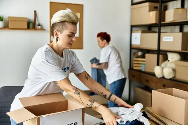 Two young women in volunteer t-shirts unpack boxes filled with items in a room, working together with enthusiasm. — Stock Photo