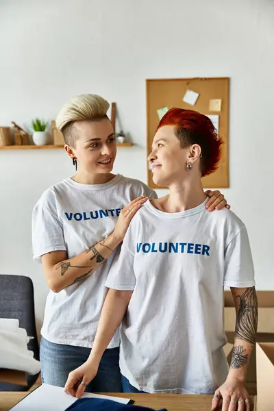 Pareja lesbiana joven en camisetas voluntarias, de pie unida en una habitación mientras hacen trabajo de caridad juntos. - foto de stock