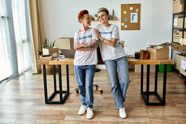 Due donne in magliette volontarie in piedi in una stanza, che lavorano insieme per una causa di beneficenza con un senso di unità e scopo. — Foto stock