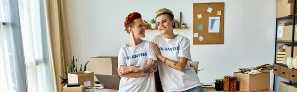 Two young women in volunteer t-shirts standing side by side in a room, united in their charitable work. — Stock Photo