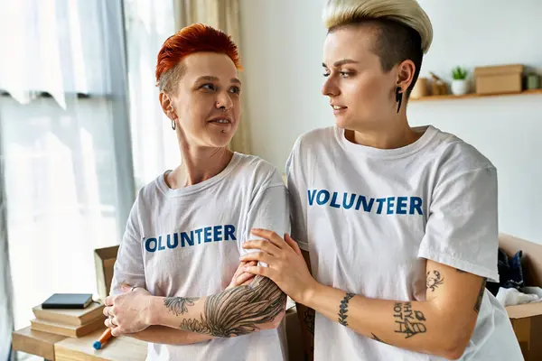 A young lesbian couple, wearing volunteer t-shirts, stands side by side, actively engaging in charity work. — Foto stock