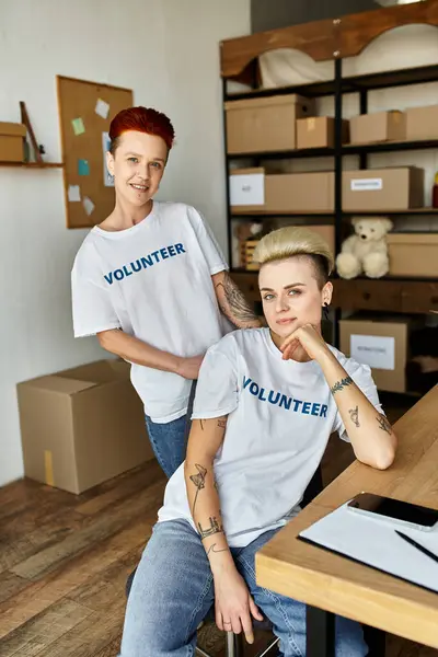 Women both wearing volunteer t-shirts, sit at a table engaged in charity work together. — Stock Photo