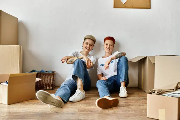 Two women, volunteers in charity shirts, sit amidst moving boxes, sharing a moment of connection and unity. — Stock Photo