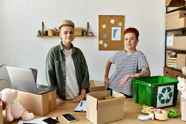 Joven pareja lesbiana está junto a una mesa llena de cajas, trabajando juntos por una causa benéfica. - foto de stock