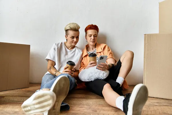 A young lesbian couple sits on the ground near boxes at a volunteer center, focused on their cell phones. — Stock Photo