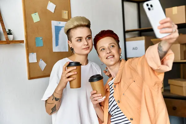 Deux jeunes femmes, un couple de lesbiennes, posent joyeusement avec une tasse de café dans les mains tout en prenant un selfie dans un centre de bénévolat. — Photo de stock