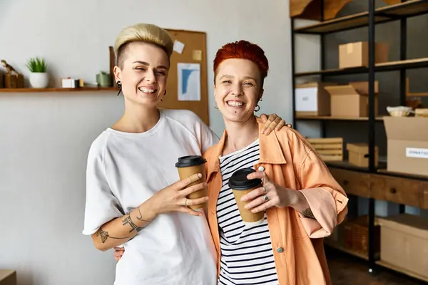A young lesbian couple standing side by side, holding coffee cups in a volunteer center surrounded by charity boxes. — Stock Photo