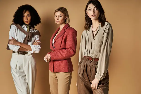 Three young, multicultural women in stylish attire stand united against a beige background, symbolizing friendship and diversity. — Stock Photo