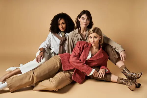 Three young, stylish, multicultural women sitting on the ground, posing for a photograph against a beige background. — Stock Photo