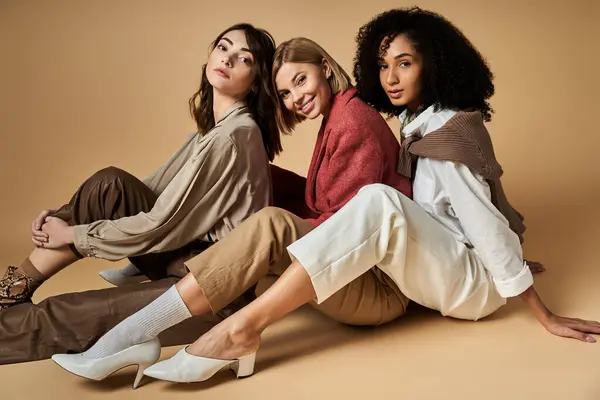 Tre giovani, donne alla moda di culture diverse siedono a terra, in posa per una foto con sfondo beige. — Foto stock