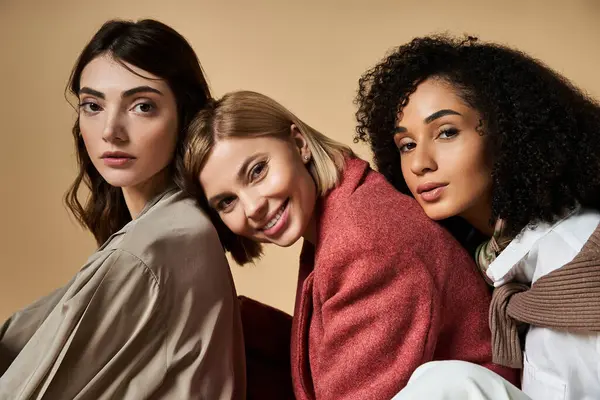 Three young, beautiful, multicultural women in stylish attire embracing their diversity while sitting together on a beige background. — Stock Photo