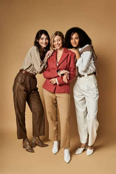 Three young, beautiful women of diverse cultures and backgrounds stand together in stylish attire on a beige background. — Stock Photo