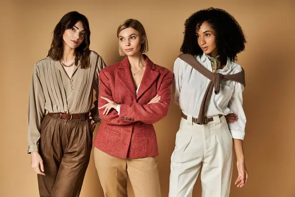 Three young, beautiful women of diverse cultural backgrounds pose together in stylish attire on a neutral beige backdrop. — Stock Photo