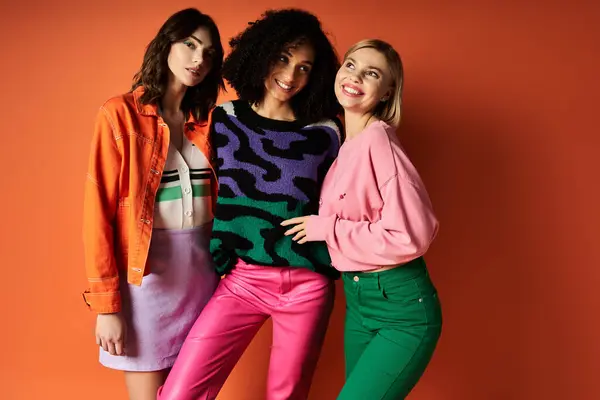 Three young, beautiful women in stylish, vibrant clothes stand together in front of an orange wall. — Stock Photo