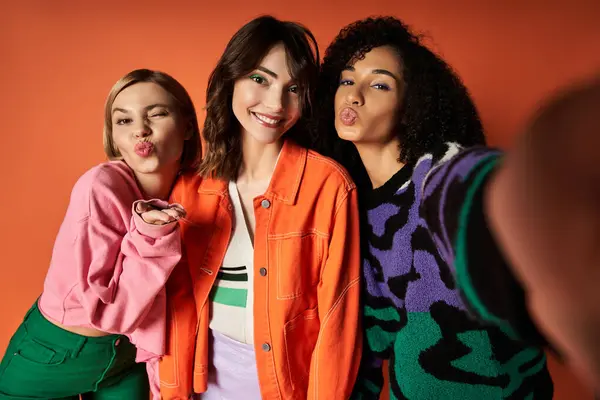 Três mulheres jovens e elegantes em roupas vibrantes estão unidas, mostrando amizade e diversidade cultural em um pano de fundo laranja. — Fotografia de Stock