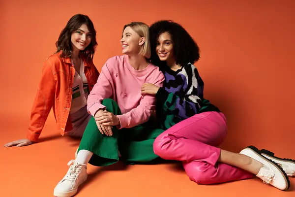 Três jovens mulheres em roupas vibrantes, sentadas no chão, sorrindo e posando para uma foto em um fundo laranja. — Fotografia de Stock