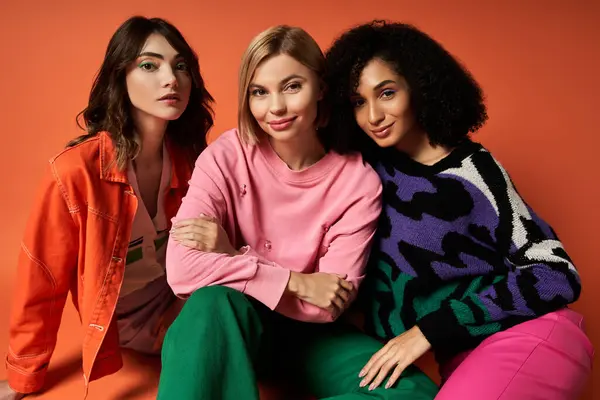 Three beautiful multicultural women in stylish attire pose for a portrait together against an orange backdrop. — Stock Photo