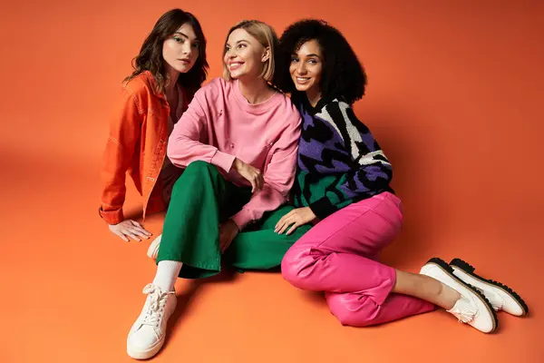 Three young, multicultural women in stylish clothes sit on the ground, posing for a fun picture against an orange backdrop. — Stock Photo