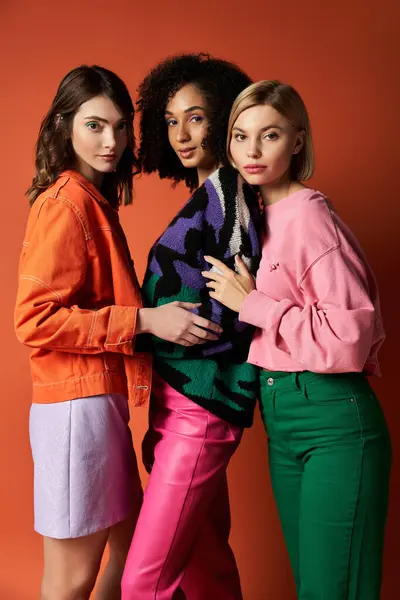 Three young, stylish women of different cultures standing together on an orange background, showcasing friendship and diversity. — Stock Photo