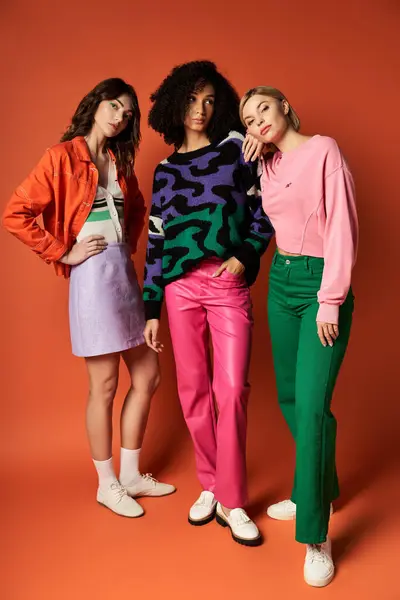 Tre giovani donne in abiti vivaci alla moda si distinguono in unità, mettendo in mostra la diversità nelle culture su uno sfondo arancione. — Foto stock