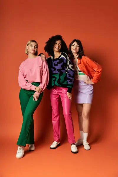 Three young beautiful multicultural women in stylish vibrant clothes stand united on an orange background. — Stock Photo
