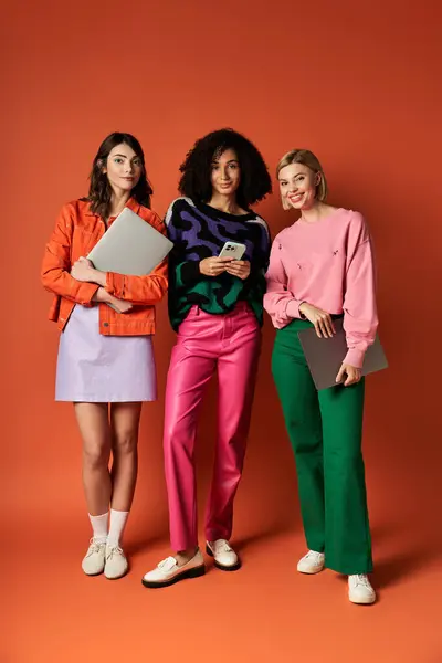Young beautiful women in vibrant clothing stand together before an orange backdrop, showcasing friendship and cultural diversity. — Stock Photo