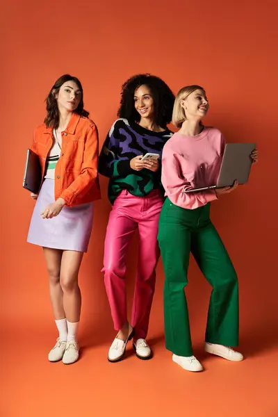 Drei junge Frauen unterschiedlicher kultureller Herkunft posieren in stylischer Kleidung vor orangefarbenem Hintergrund. — Stockfoto
